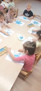 Kilkoro dzieci siedzi przy stoliku malując palcami śnieżynkę niebieską farbą 