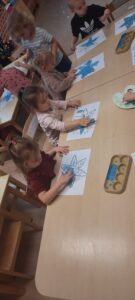 Grupa dzieci siedzi przy stoliku malując palcami śnieżynki niebieską farbą 