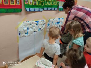 Grupa dzieci stoi naprzeciwko ściany na której jest kartka z watą kosmetyczną oraz niebieską bibułą 