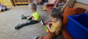 Dwóch chłopców siedzi na dywanie, każdy ma 6 kolorowych klocków 