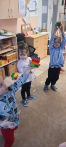 Dzieci stoją na dywanie, w dłoniach trzymają kostkę z kolorowych klocków 