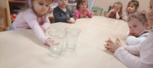 Grupa dzieci siedzi przy stoliku, przed nimi dwa kubki plastikowe z wodą 