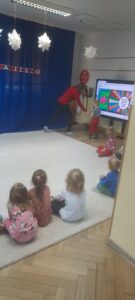 Grupa dzieci siedzi na dywanie patrząc w stronę tablicy multimedialnej 