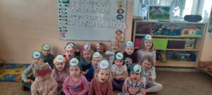 Grupa dzieci siedzi na dywanie, każde ma na głowie opaskę z dinozaurem, za nimi na tablicy napis "dzień dinozaura" 