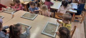 Grupa dzieci siedzi przy stoliku, w dłoniach mają pędzelki, przed nimi tacki z obrazkami przykrytymi piaskiem 