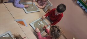 Grupa dzieci siedzi przy stoliku, w dłoniach mają pędzelki, przed nimi tacki z obrazkami przykrytymi piaskiem 
