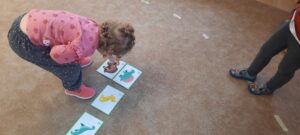 Dziewczynka stoi na dywanie kładąc szkielet dinozaura pod obrazkiem dinozaura 