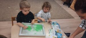 Dwoje dzieci siedzi przy stoliku, przed nimi tacka z kolorową masą