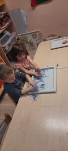 Dwoje dzieci siedzi przy stoliku, przed nimi tacka z kolorową masą