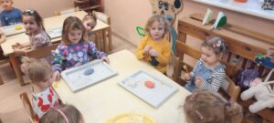 Grupa dzieci siedzi przy stoliku, przed nimi tacki z jajkami z kolorowej masy 