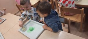 Dwoje dzieci siedzi przy stoliku, w dłoniach trzymając plastikowe łyżeczki, przed nimi plastikowa tacka z jajkiem z kolorowej masy i miseczka z wodą 