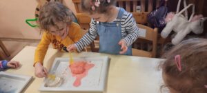Dwoje dzieci siedzi przy stoliku, w dłoniach trzymając plastikowe łyżeczki, przed nimi plastikowa tacka z jajkiem z kolorowej masy i miseczka z wodą 