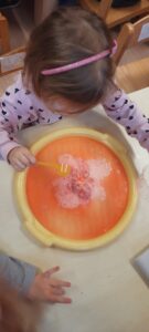 Dziewczynka siedzi przy stoliku, w dłoniach trzymając plastikowe łyżeczki, przed nią plastikowa tacka z jajkiem z kolorowej masy i miseczka z wodą 