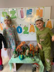 W tle napis " Dzień Dinozaura". Dwoje dzieci stoi obok stolika na którym znajdują się figurki dinozaurów