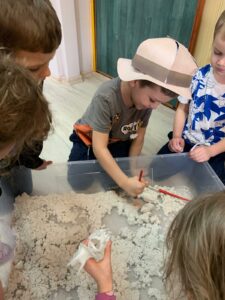 Kilkoro dzieci bawi się w paleontologów, odkopując plastikowy szkielet dinozaura ukryty pod piaskiem kinetycznym w przezroczystym pudełku.