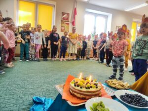 Grupa dzieci śpiewająca "sto lat". Obok tort urodzinowy owocami.