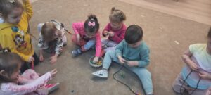 Grupa dzieci siedzi na dywanie nawlekajac kolorowe koraliki na sznurki 