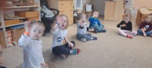 Dzieci siedzą na dywanie podnosząc rękę do góry, na nadgarstkach mają frotki 