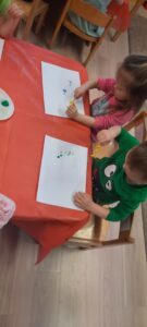 Dzieci siedzą przy stoliku malując farbami przy użyciu sztucznych kwiatów 