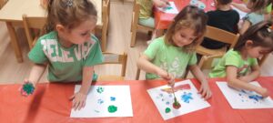 Dzieci siedzą przy stoliku malując farbami przy użyciu sztucznych kwiatów 