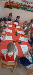 Dzieci siedzą przy stoliku na którym znajdują się Pojemniki z kolorową wodą oraz obrazki pisanek