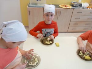 troje dzieci siedzi przy stole i kroi za pomocą noża na małe kawałki banana