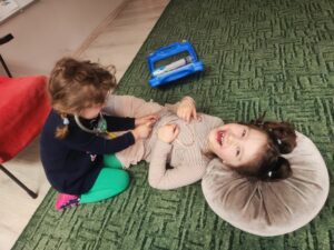 Dwie dziewczynki z przyborami lekarskimi. Jedna dziewczynka osłuchuje drugą zabawkowym stetoskopem.