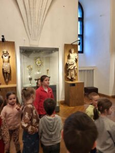 kilkoro dzieci stoi w sali muzealnej z pomnikami przedstawiającymi postacie świętych
