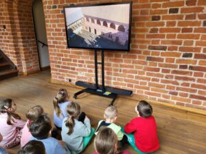 grupa dzieci siedzi przed ekranem na którym wyświetlany jest film o historii powstania zamku