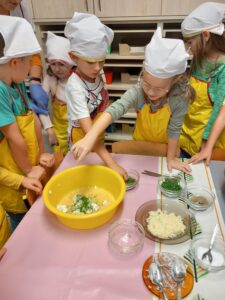 kilkoro dzieci ubranych w kolorowe fartuszki i w białych chusteczkach na głowie stoją przed stolikiem, na którym znajduje się miska z ciastem cukiniowym, dziewczynka wsypuje do ciasta szczypiorek