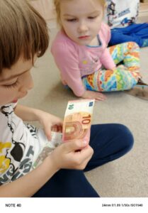 Chłopiec siedząc na dywanie trzyma w dłoni banknot 10 euro. Dziewczynka przypatruje się walucie Europy.