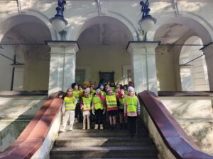 Gruoa dzieci w odblaskowych kamizelkach stoi przed wejściem do budynku.