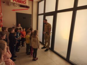 Dzieci na sali edukacyjnej, wysłuchują instrukcji strażaka.