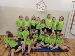 Grupa dzieci siedząca na materacu, ubrana w strój sportowy trzymająca logo przedszkola.