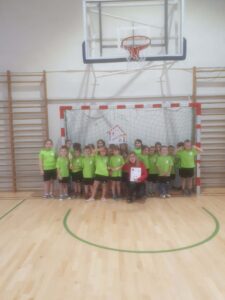 Grupa dzieci stojąca na sali gimnastycznej. W tle wiszący kosz do koszykówki