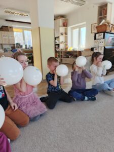 Grupa dzieci siedzi na dywanie i dmucha białe balony.
